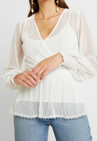 Blusa bianca in pizzo con design avvolgente, maniche lunghe in tessuto trasparente, tessuto testurizzato e orlo arricciato. Abbinata a jeans di denim azzurro chiaro.