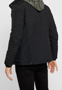 Person som bär en svart jacka med vadderade armbågar och en kontrasterande grå huva, sedd bakifrån stående mot en neutral bakgrund.