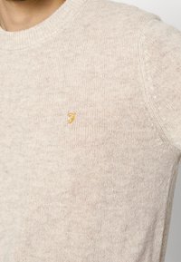 Sweater en tricot beige avec un col rond, comportant un petit logo 'F' jaune sur la poitrine et une finition en tissu texturé et doux.