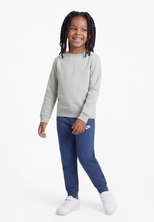 Besikalbėjęs vaikas, apsiavęs pilką džemperį, mėlynas Nike sportines kelnes ir baltus sportinius batelius, stovintis prieš baltą foną.