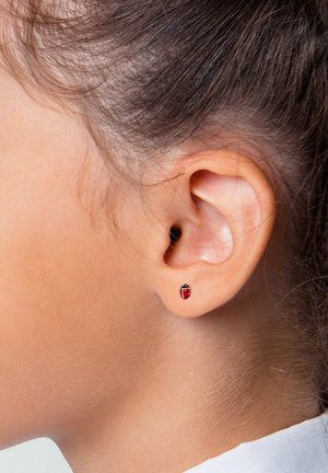 Nahaufnahme eines Ohres einer Person mit zurückgebundenem braunem Haar, das einen kleinen roten Ohrring in Form eines Marienkäfers trägt.