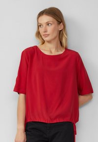 Blouse rouge à manches courtes en tissu léger avec un col rond et un ourlet élastique froncé. Texture lisse avec une coupe décontractée.