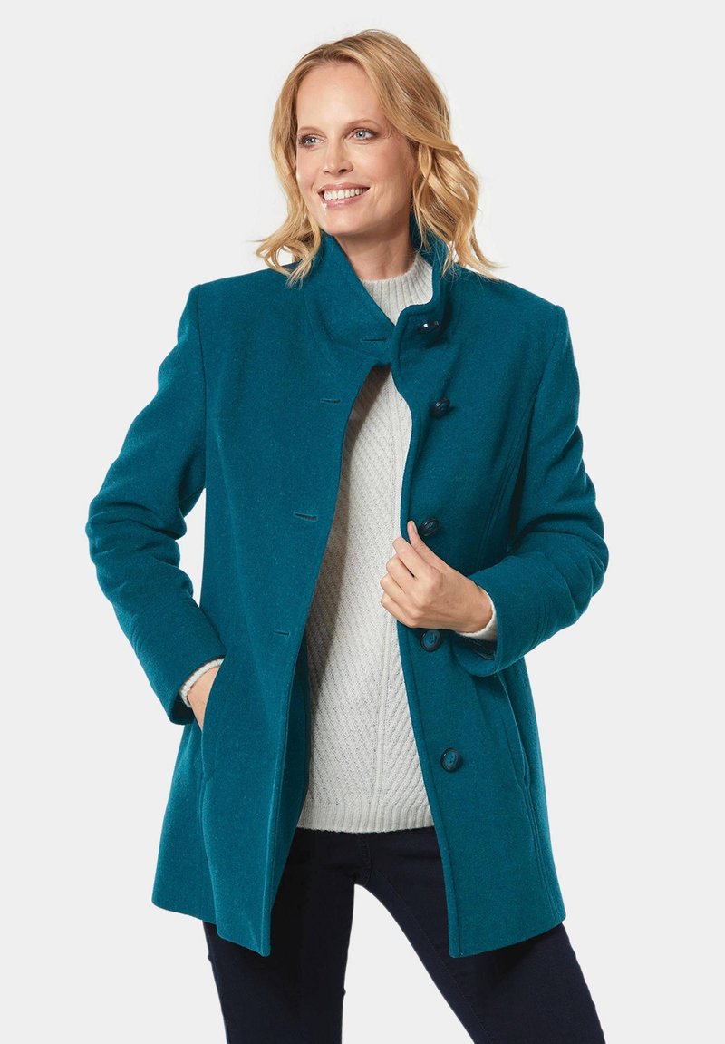Türkisfarbener Wollmischmantel mit hohem Kragen, ausgestattet mit einer einzelnen Knopfreihe und Seitentaschen, kombiniert mit einem leichten Pullover darunter.