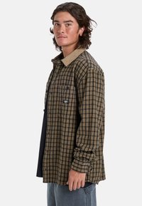 Camisa de franela a cuadros marrón y negra con un cuello de pana color beige, bolsillo en el pecho y cierre de botones. Se lleva sobre una camiseta negra.