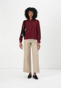 Cămașă burgundy cu mânecă lungă, cu nasturi, dintr-un material texturat, asortată cu pantaloni bej cu croială largă și mocasini negri. Geantă neagră pe umăr.