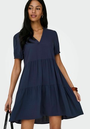 Ženska v ohlapni dolgoročni temno modri obleki s kratkimi napihnjenimi rokavi, drži črno torbico v eni roki in nosi rjavo zapestnico na drugi.