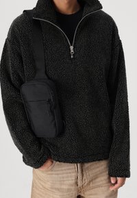 Sudadera de forro polar negra con cuello alto y cierre de cremallera, sobre pantalones beige claros, acompañada de un bolso cruzado compacto negro.