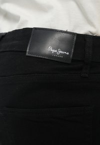 Pantalones vaqueros de mezclilla negra con una etiqueta de cuero visible que presenta "Pepe Jeans London" en escritura blanca. Incluye un bolsillo trasero y detalles sutiles de costura.