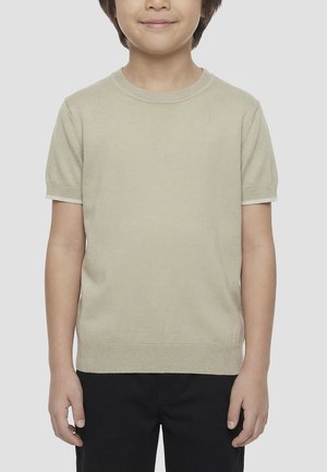 Dijete u bež kratkoj pletenoj majici i crnim hlačama, stoji s opuštenim rukama na jednostavnoj svijetlosivoj pozadini.