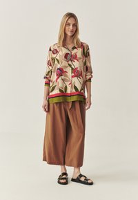 Vrouw in een bloemenprint lange mouwen shirt, brede bruine cropped broek en zwarte sandalen met open tenen, staand tegen een effen beige achtergrond.