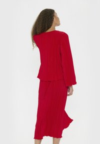 Donna con capelli ricci che indossa un top lungo a maniche e a pieghe di un rosso brillante e pantaloni larghi abbinati, di spalle su uno sfondo bianco.