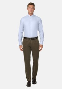 Helles blaues Langarm-Hemd mit Button-Down-Kragen, kombiniert mit olivgrünen Cordhosen und schwarzen Schuhen, stehend vor einem neutralen Hintergrund.