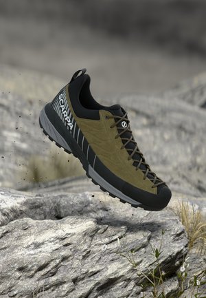 Donkergroene en zwarte wandelschoen met "Scarpa"-logo in de lucht boven rotsachtig terrein met enkele kleine planten.