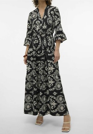 Frau trägt ein schwarzes und weißes, blumiges Maxikleid mit drei Viertelärmeln, einem gebundenen Taillengürtel und beigen offenen Sandalen.