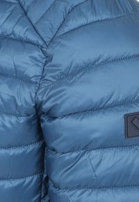 Gewatteerde blauwe jas met een elegante, gladde textuur en een ruitpatroon, voorzien van een klein zwart logo-opdruk op de schouder.