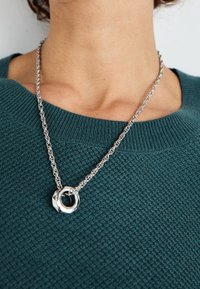 Collier en chaîne argentée avec un pendentif circulaire porté sur un pull texturé bleu sarcelle foncé, autour du cou et du haut de la poitrine d'une personne.