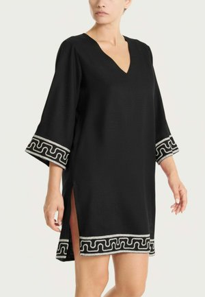 Donna che indossa un vestito nero con scollo a V, maniche larghe e bordatura decorativa argentata su polsini e orlo, con spacchi laterali.