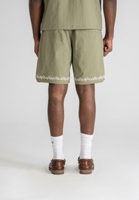 Olivenfargede shorts med vertikale striper, med hvit blomsterbroderi langs kanten. Brukt med hvite sokker og brune sko.