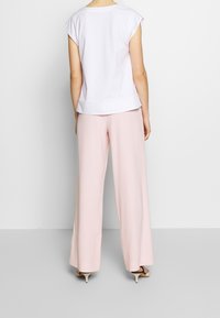 Mujer de pie de espaldas a la cámara, que lleva una blusa blanca de manga corta, pantalones anchos de color rosa claro y sandalias de tacón bajo beige.