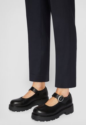 Chaussures Mary Jane en cuir noir avec semelles épaisses à crampons et brides à boucle ronde argentée, portées avec un pantalon court foncé.