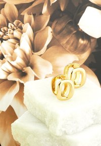 Guldörhängen med en avlång oval design som vilar på vita stenar, mot en mjukt suddig blomsterbakgrund.