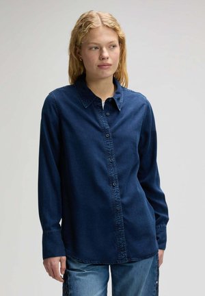 Junge Frau blickt seitlich, trägt ein langärmliges, dunkelblaues Jeanshemd mit Knöpfen und hellblaue Jeans vor einfachem Hintergrund.