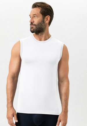 Man met baard en kort haar, gekleed in een effen wit mouwloos shirt, kijkt naar links tegen een neutrale achtergrond.