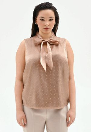 Donna che indossa una blusa beige senza maniche con piccoli pois bianchi e fiocco annodato al collo, abbinata a pantaloni chiari, in piedi e rivolta verso l'osservatore.