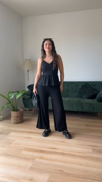 Top peplum preto combinado com calças largas pregueadas pretas. A modelo segura uma bolsa de mão tecida preta e usa sapatos plataforma chunk claros pretos.