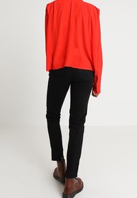 Blouse rouge à manches longues avec une coupe décontractée et des poignets boutonnés, associée à un jean skinny noir avec un accent à fermeture éclair sur le côté et des bottines brunes.