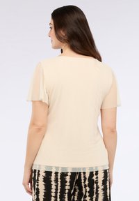 Donna con lunghi capelli castani che indossa una maglietta beige a maniche corte e pantaloni a fantasia bianco e nero, di spalle su uno sfondo bianco semplice.