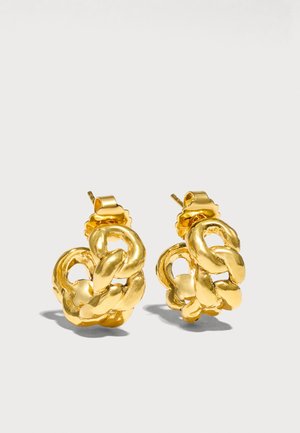Paire de boucles d’oreilles à clou en métal doré brillant avec maillons de chaîne, dotées de fermoirs à tige, projetant de douces ombres sur un fond clair uni.