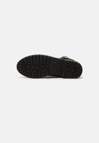 Botte noire avec une semelle en caoutchouc texturée, présentant des crampons profonds et un motif rectangulaire. Les accents incluent une boucle métallique.