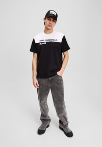Juoda ir balta marškinėliai su užrašu "KARL LAGERFELD JEANS", derinami su nusidėvėjusiais pilkais džinsais ir juodais batais. Paprastas, kasdieniškas dizainas su kontrastingomis spalvomis.