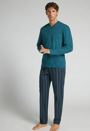 Junger Mann steht barfuß, trägt ein türkisfarbenes Langarm-V-Neck-Shirt mit Brusttasche und marineblaue gestreifte Pyjamahosen.