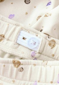 Tissu beige clair avec des pois dorés et violets. Comprend une taille élastique blanche avec une fermeture à bouton et des détails cousus.