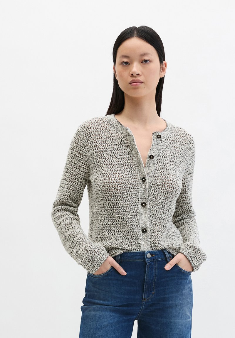 Femme aux cheveux noirs lisses portant un cardigan en maille texturée gris clair avec des boutons noirs et un jean en denim bleu, les mains dans les poches.