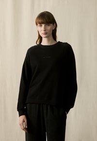 Sudadera negra con cuello redondo, mangas raglán y dobladillo recto. Presenta un sutil logo en la parte delantera central. Textura de tela suave.