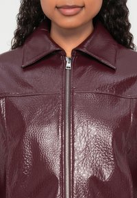 Burgundska jakna iz umetnega usnja z visokim ovratnikom, sprednjim zadrgo in teksturirano površino. Ima vidne šive in dva prsna žepa.