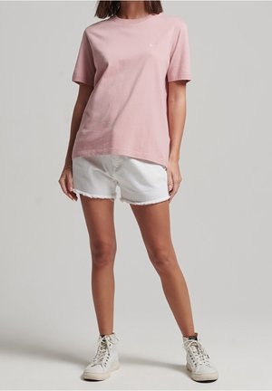 Personne portant un t-shirt rose clair à manches courtes, un short en jean blanc et des baskets blanches montantes, debout devant un fond neutre.