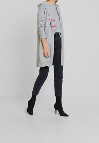 Grå lång cardigan med huva, bärs över en ljusgrå tröja med en rosa "C"-logotyp, i kombination med svarta skinny jeans och svarta klackiga ankelstövlar.