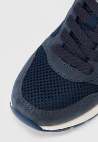 Σκούρο μπλε αθλητικό παπούτσι με συνδυασμό διχτυωτού υφάσματος και σουέντ, με επένδυση στον πάτο και ανάγλυφο σχέδιο με κορδόνια στο πάνω μέρος.