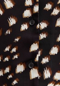 Zwarte stof met een abstracte luipaardprint in bruin en crème. Voorzien van zwarte knopen en een zachte textuur. Close-up van de detail van het kledingstuk.