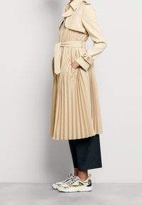 Beige plisserad trenchcoat med bälte i midjan och sidofickor, matchad med svarta beskurna byxor och mönstrade sneakers. Slät tygtextur.