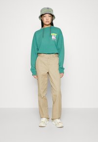 Camicetta verde a maniche lunghe con un design grafico, pantaloni beige e un cappello a secchiello a righe. Le sneakers bianche completano l'outfit.