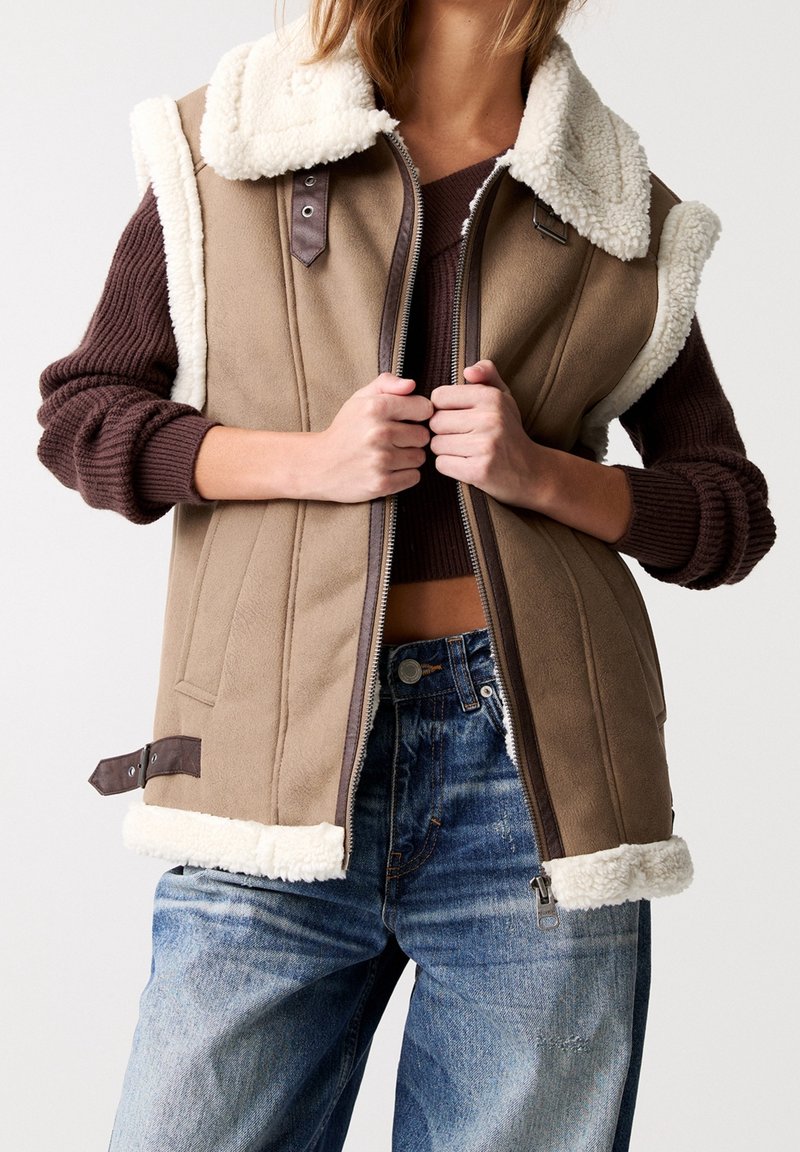 Gilet marrone senza maniche con colletto in sherpa crema, zip frontale e dettagli in pelle. Indossato sopra un maglione a maglia e jeans. Tessuto testurizzato.