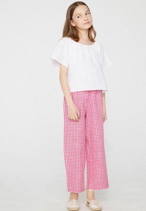 Fille aux longs cheveux portant un chemisier blanc à manches courtes, un pantalon large à carreaux rose et blanc, et des espadrilles beiges, debout et regardant sur le côté.