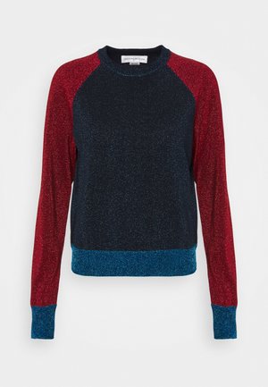 Henkilö, joka pukeutunut kimaltavaan punaisiin ja sinisiin pitkähihainen yläosaan, löysiin sinisiin farkkuihin, hopeisiin kimaltaviin kenkiin ja pitää kädessään punaista clutch-laukkua.