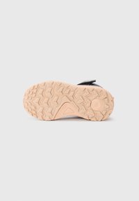 Semelle de chaussure en caoutchouc avec un motif de grip texturé en beige, mettant en valeur des accents marron clair et un dessus en tissu noir. Une bande Velcro est visible en haut.