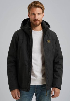 Schwarze Jacke mit Kapuze, Reißverschluss vorne und einer Brusttasche. Verfügt über ein gestepptes Design und strukturierter Stoff. Kombiniert mit einem hellgrauen Hemd und blauen Jeans.